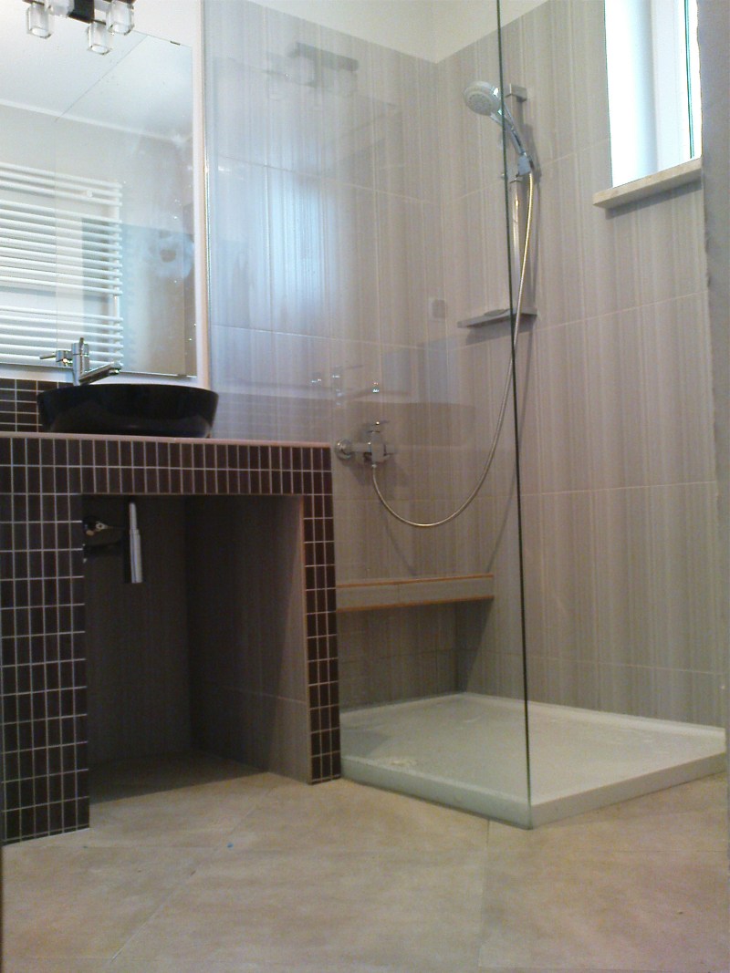 Nowoczesna łazienka z czarną, okrągłą umywalką nablatową, kabiną prysznicową ze szklaną ścianą i szarymi płytkami ściennymi o subtelnej fakturze.